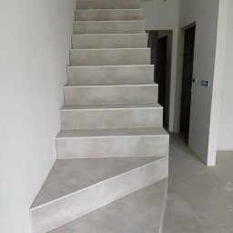 SERHII KOROLOV - Schody wyłożone szarymi płytkami, widok z dołu, surowy stan wykończenia wnętrza, ściany pomalowane na biało, widoczne puszki elektryczne bez osprzętu.