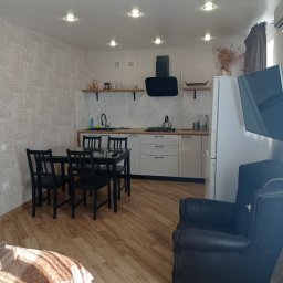 SERHII KOROLOV - Jasne wnętrze mieszkania z drewnianą podłogą, widoczny fragment sypialni z beżową narzutą, czarny stół z krzesłami oraz aneks kuchenny z białymi szafkami i marmurowym blatem.