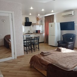 SERHII KOROLOV - Jasne wnętrze mieszkania typu studio z drewnopodobną podłogą, tapetowanymi ścianami, aneksem kuchennym, czarnymi krzesłami przy stole i łóżkiem z brązową narzutą oraz złożonymi ręcznikami