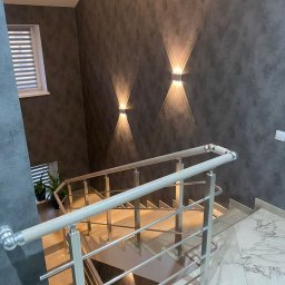 SERHII KOROLOV - Klatka schodowa z drewniano-metalowymi balustradami, ściany w ciemnym odcieniu z dekoracyjnymi lampami ściennymi rzucającymi stożkowe światło, marmurowe płytki podłogowe, widok z góry.