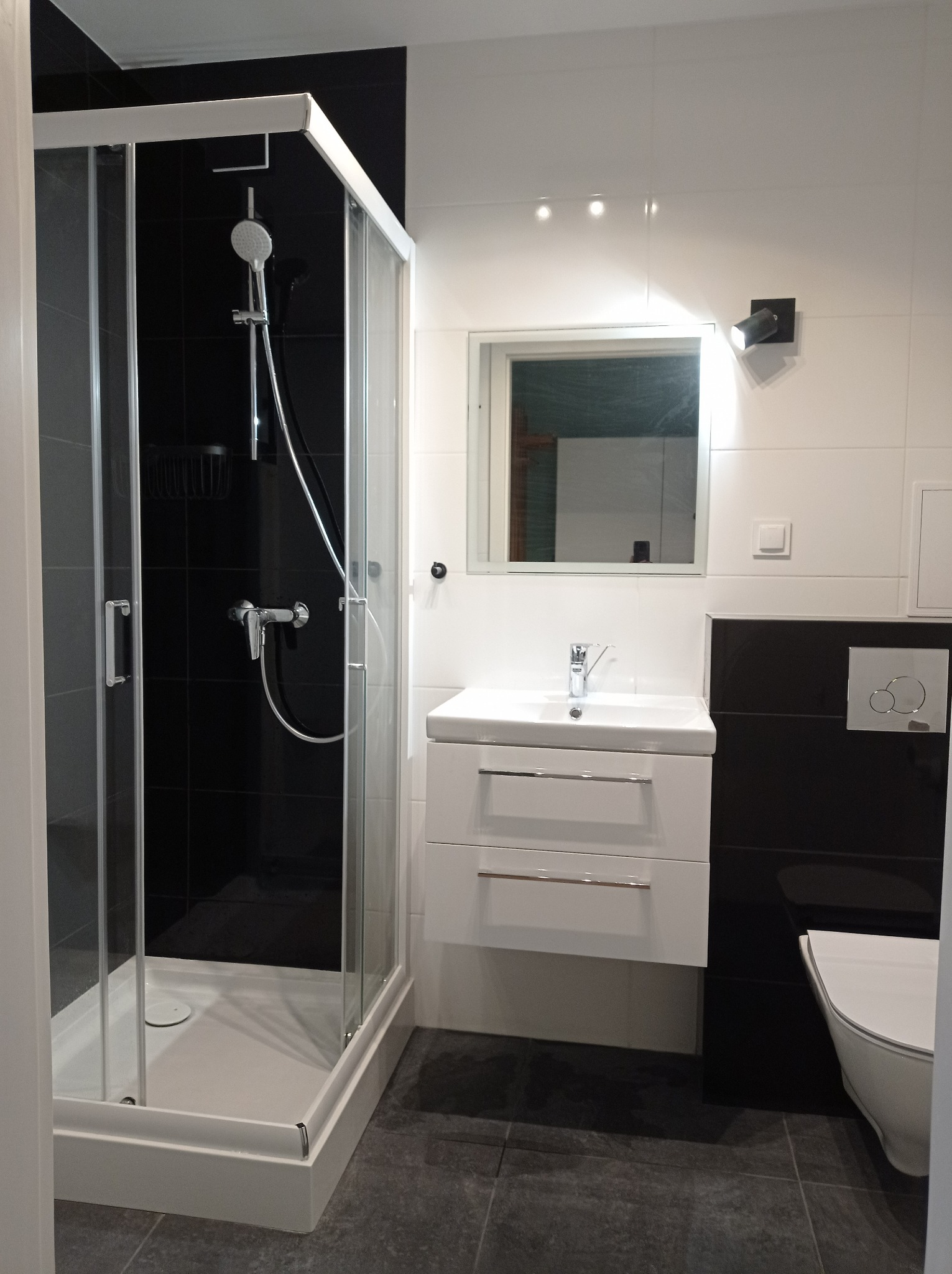 Nowoczesna łazienka z czarno-białymi płytkami, kabiną prysznicową z chromowanym wykończeniem, białą umywalką z szafką i lustrem, oraz toaletą w zabudowie.