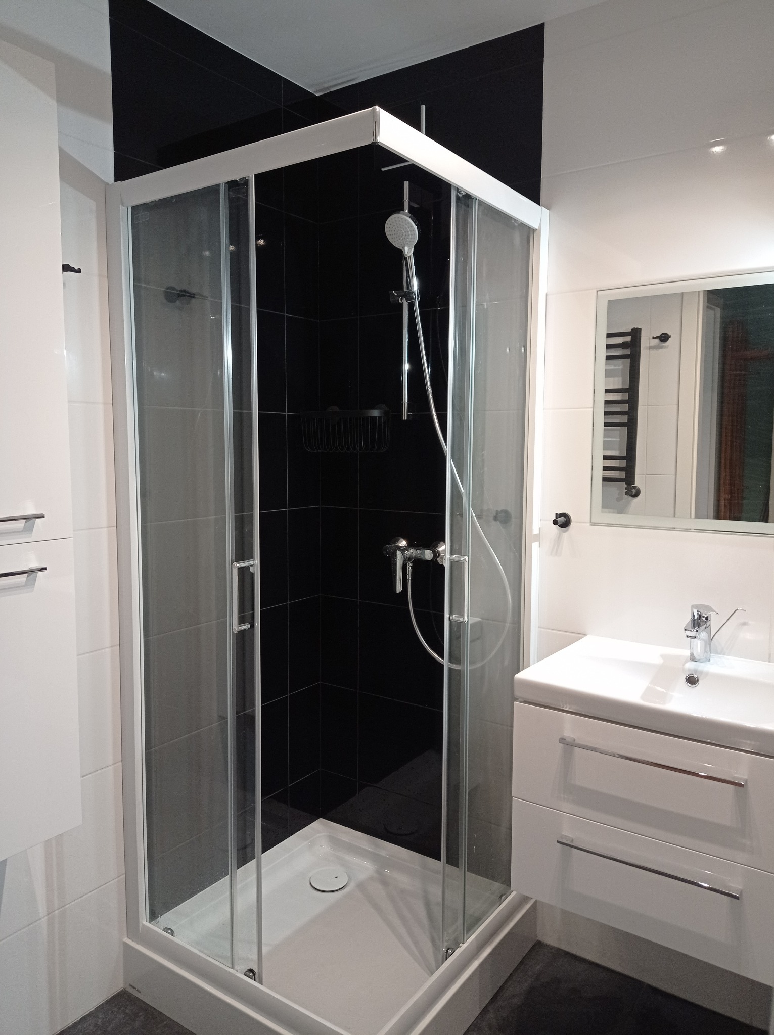 Nowoczesna łazienka z kabiną prysznicową z przesuwnymi drzwiami, czarnymi płytkami wewnątrz kabiny i białą umywalką z szafką.