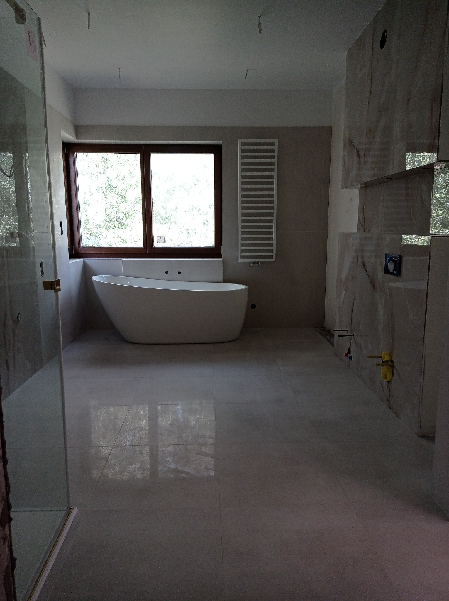 Wnętrze łazienki w trakcie wykańczania, widoczna wolnostojąca wanna przy oknie, grzejnik drabinkowy na ścianie, częściowo ułożone marmurowe płytki na ścianach, instalacja wodna wystająca ze ściany.