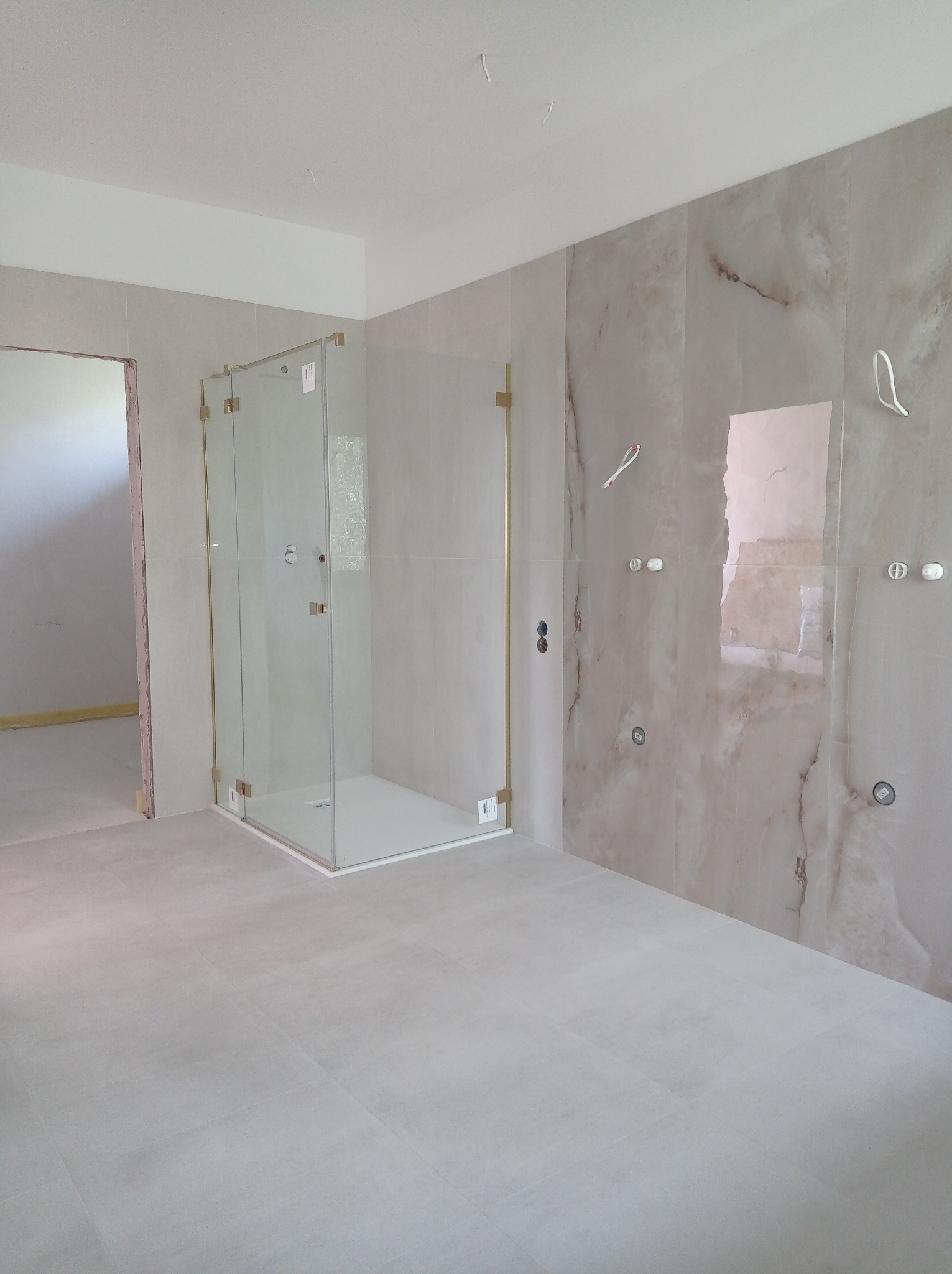 Nowoczesna łazienka w trakcie wykańczania: kabina prysznicowa ze złotymi okuciami, marmurowe płytki ścienne z instalacją elektryczną, szare płytki podłogowe i surowy otwór drzwiowy.