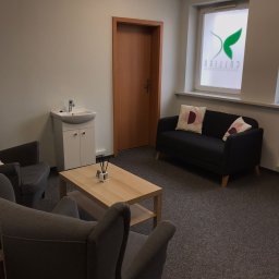 Centrum Psychiatrii i Psychoterapii COLLIGO w Bydgoszczy - Jasne wnętrze gabinetu z dwoma szarymi fotelami, ciemną sofą z poduszkami, drewnianym stolikiem kawowym z dyfuzorem zapachowym, umywalką z szafką i oknem z logo firmy.