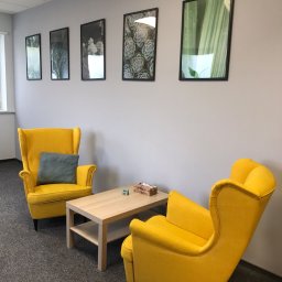 Centrum Psychiatrii i Psychoterapii COLLIGO w Bydgoszczy - Jasne, nowoczesne wnętrze gabinetu z dwoma żółtymi fotelami typu uszak, drewnianym stolikiem kawowym i serią obrazków z motywami roślinnymi na szarej ścianie.