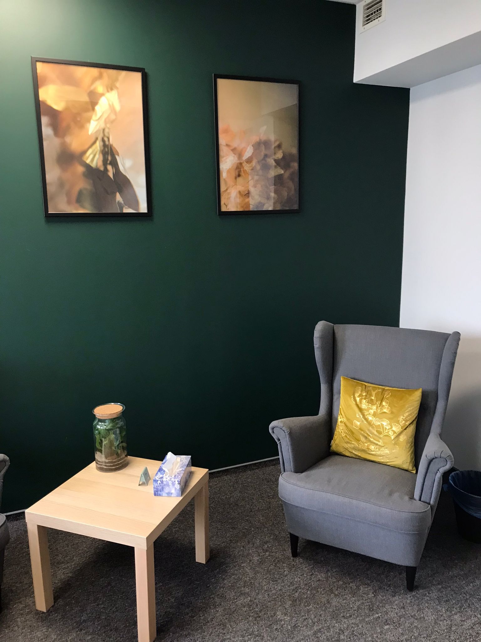 Wnętrze gabinetu z ciemnozieloną ścianą, dwoma obrazami w czarnych ramkach, jasnym stolikiem z pudełkiem chusteczek i słoikiem z roślinami, oraz szarym fotelem z żółtą poduszką.
