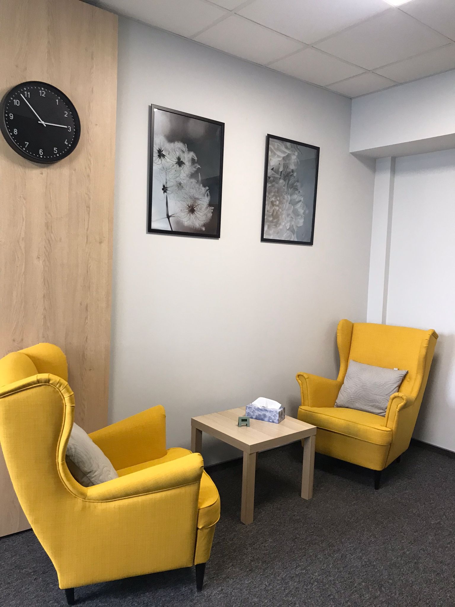 Jasne wnętrze gabinetu z dwoma żółtymi fotelami typu uszak, stolikiem z chusteczkami i zegarem na ścianie.