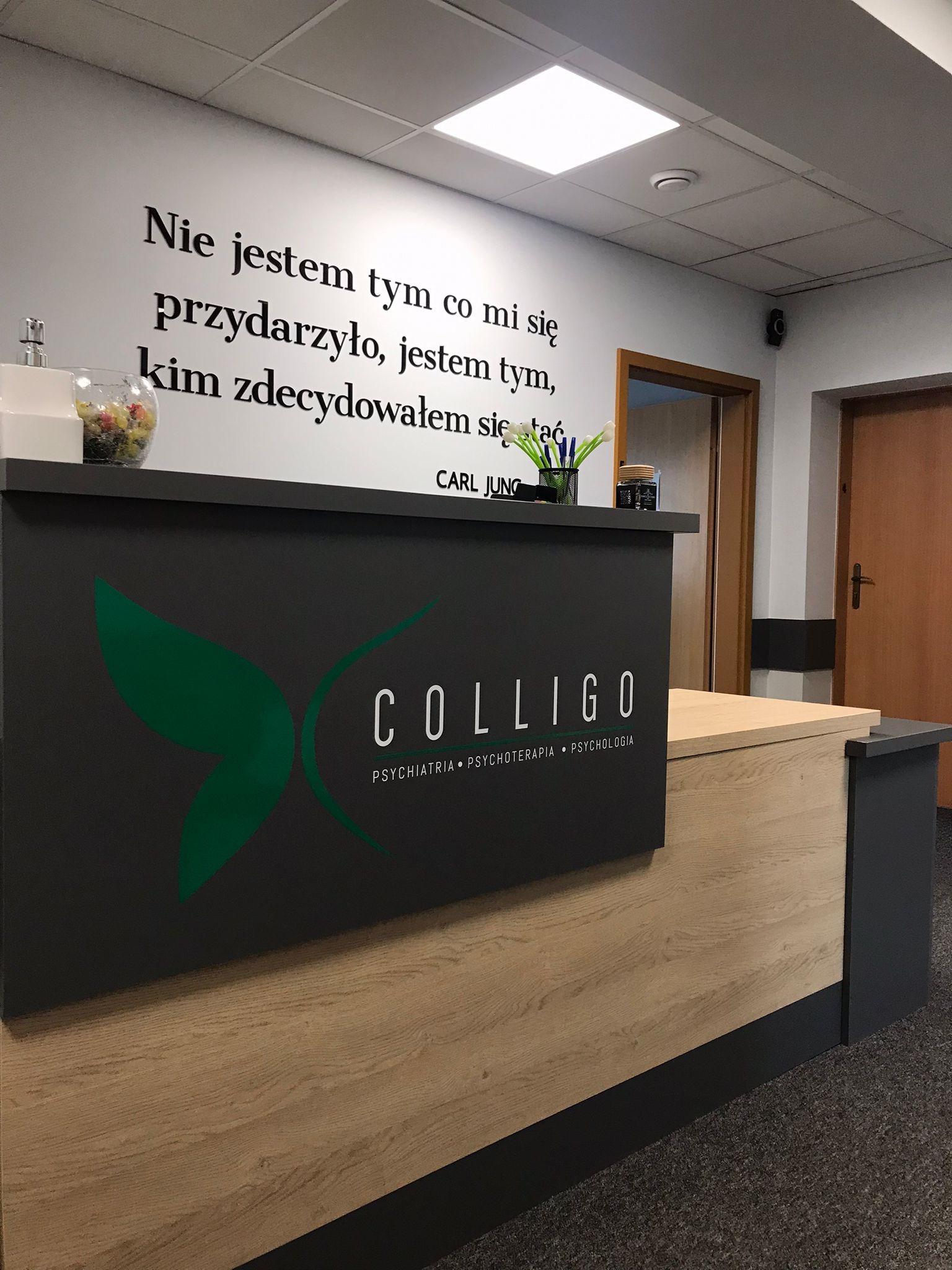 Recepcja w placówce medycznej z logo firmy COLLIGO, oferującej psychiatrię, psychoterapię i psychologię. Na ścianie cytat Carla Junga w czarnym druku.
