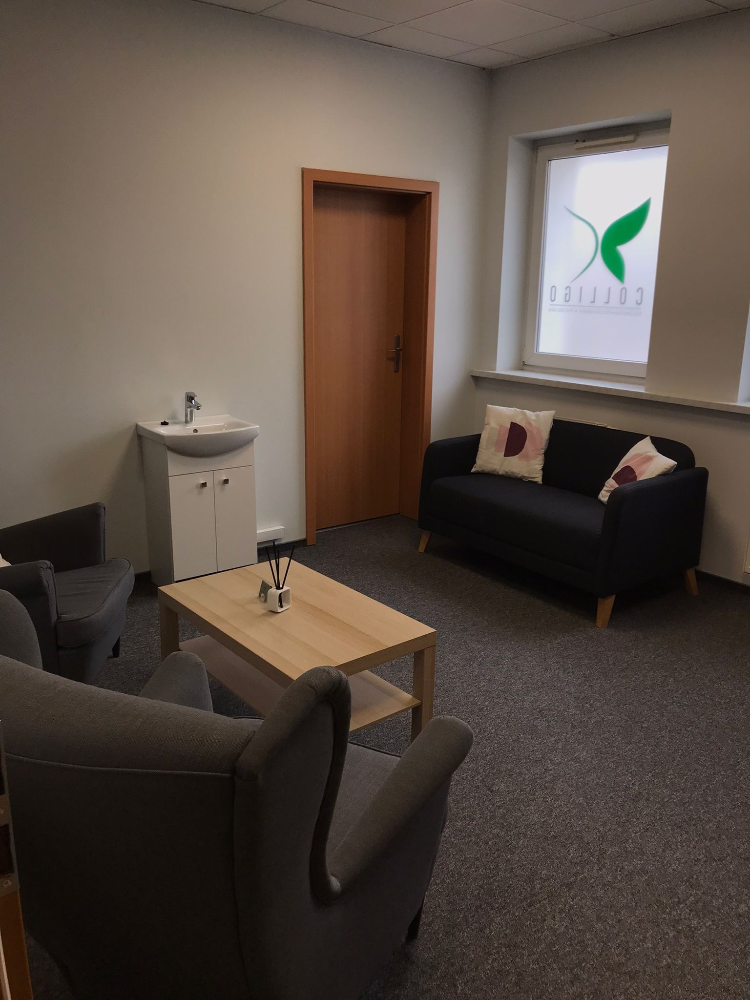 Jasne wnętrze gabinetu z dwoma szarymi fotelami, ciemną sofą z poduszkami, drewnianym stolikiem kawowym z dyfuzorem zapachowym, umywalką z szafką i oknem z logo firmy.