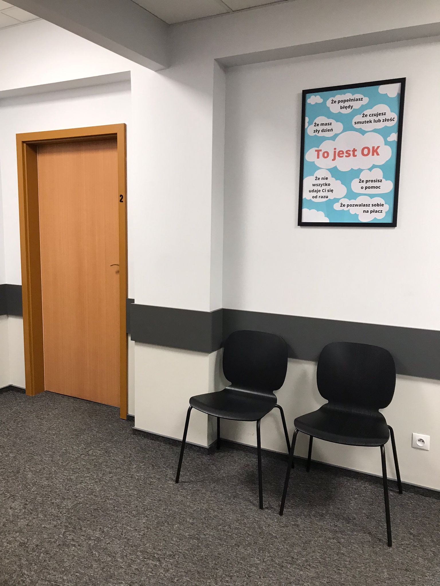 Poczekalnia z dwoma czarnymi krzesłami, otwartymi drzwiami z numerem 2 i plakatem na ścianie z hasłem 'To jest OK' i listą akceptowalnych emocji.