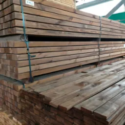 Stosy brązowych desek ułożone w paczki, zabezpieczone taśmą, składowane w magazynie z widoczną konstrukcją stalową dachu.