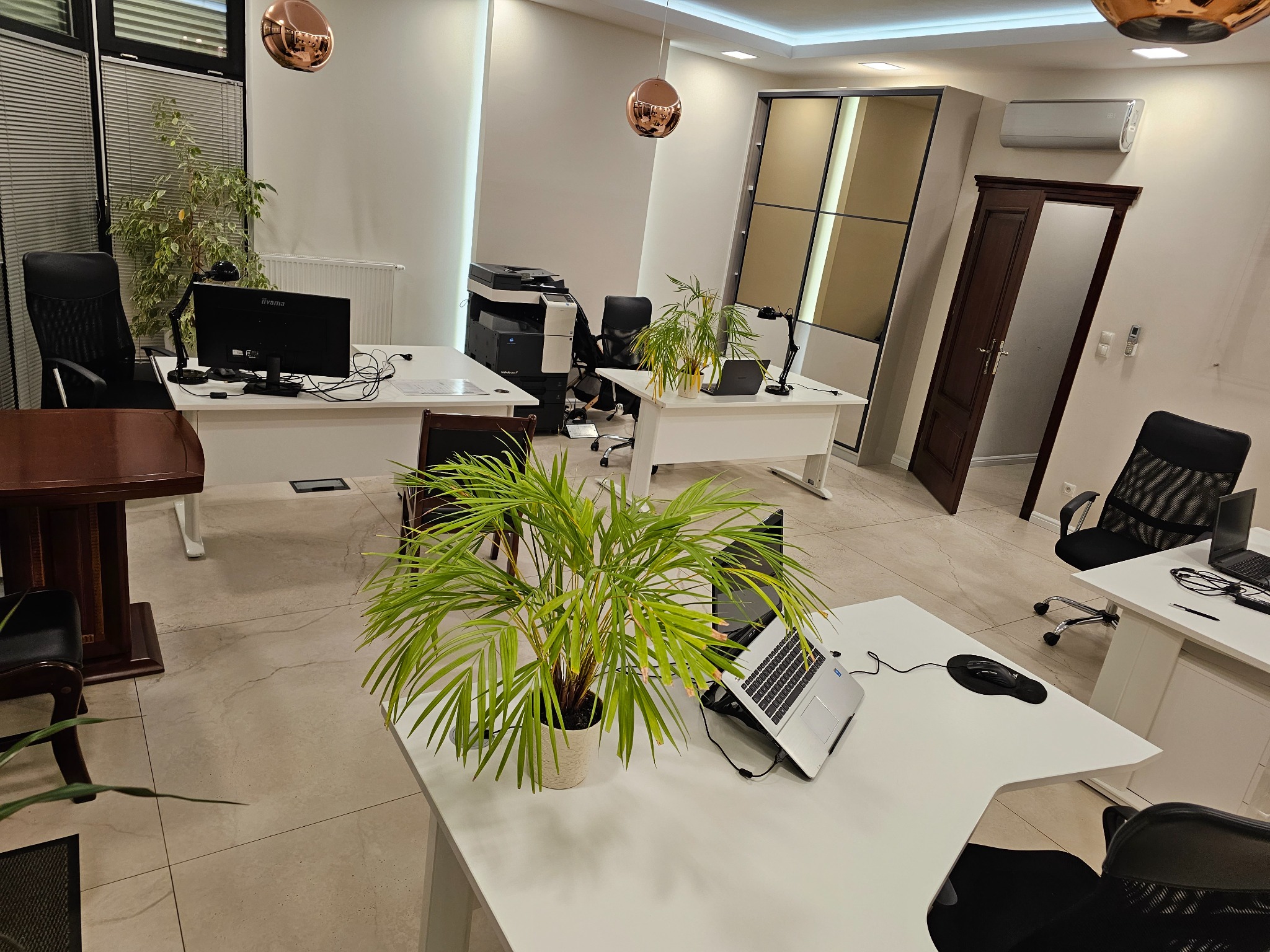 Jasne biuro z białymi biurkami, komputerami i roślinami doniczkowymi, oświetlone wiszącymi lampami w kolorze miedzi.