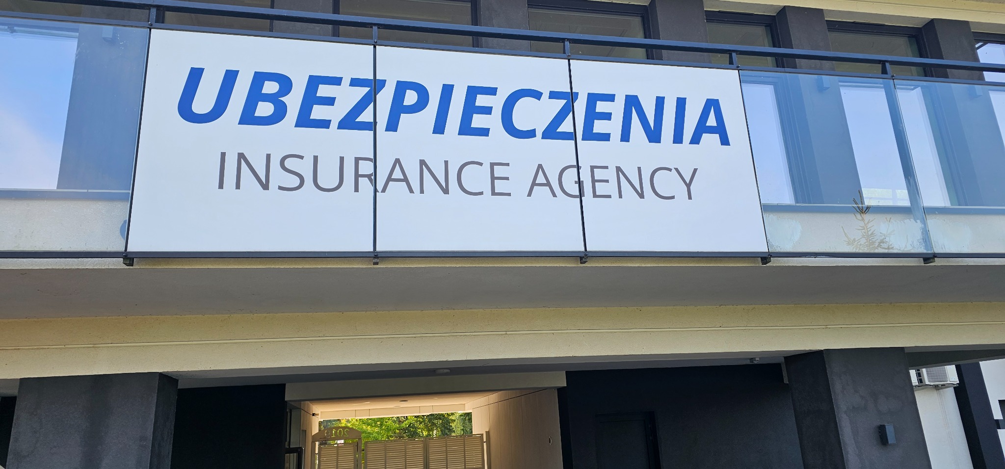 Szyld z napisem 'Ubezpieczenia Insurance Agency' nad wejściem do biura w Nowym Dworze Mazowieckim, widok z dołu, na szklanej balustradzie odbija się mała roślina.