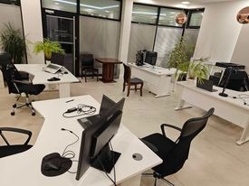 Jasne, przestronne biuro z kilkoma stanowiskami pracy. Białe biurka z komputerami i krzesłami biurowymi. Rośliny doniczkowe dodają świeżości. Widoczne okna i drzwi wejściowe.