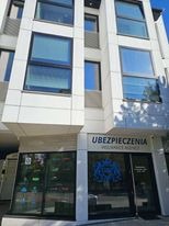 Fasada nowoczesnego budynku z biurem agencji ubezpieczeniowej na parterze, widoczne okna wystawowe z grafikami i napisem 'Ubezpieczenia Insurance Agency' nad wejściem.