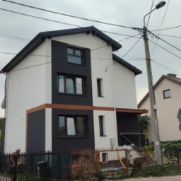 Dwukondygnacyjny dom w Białymstoku po częściowym malowaniu elewacji na biało i czarno, z widoczną pomarańczową linią oddzielającą kolory, prace porządkowe na posesji w toku.