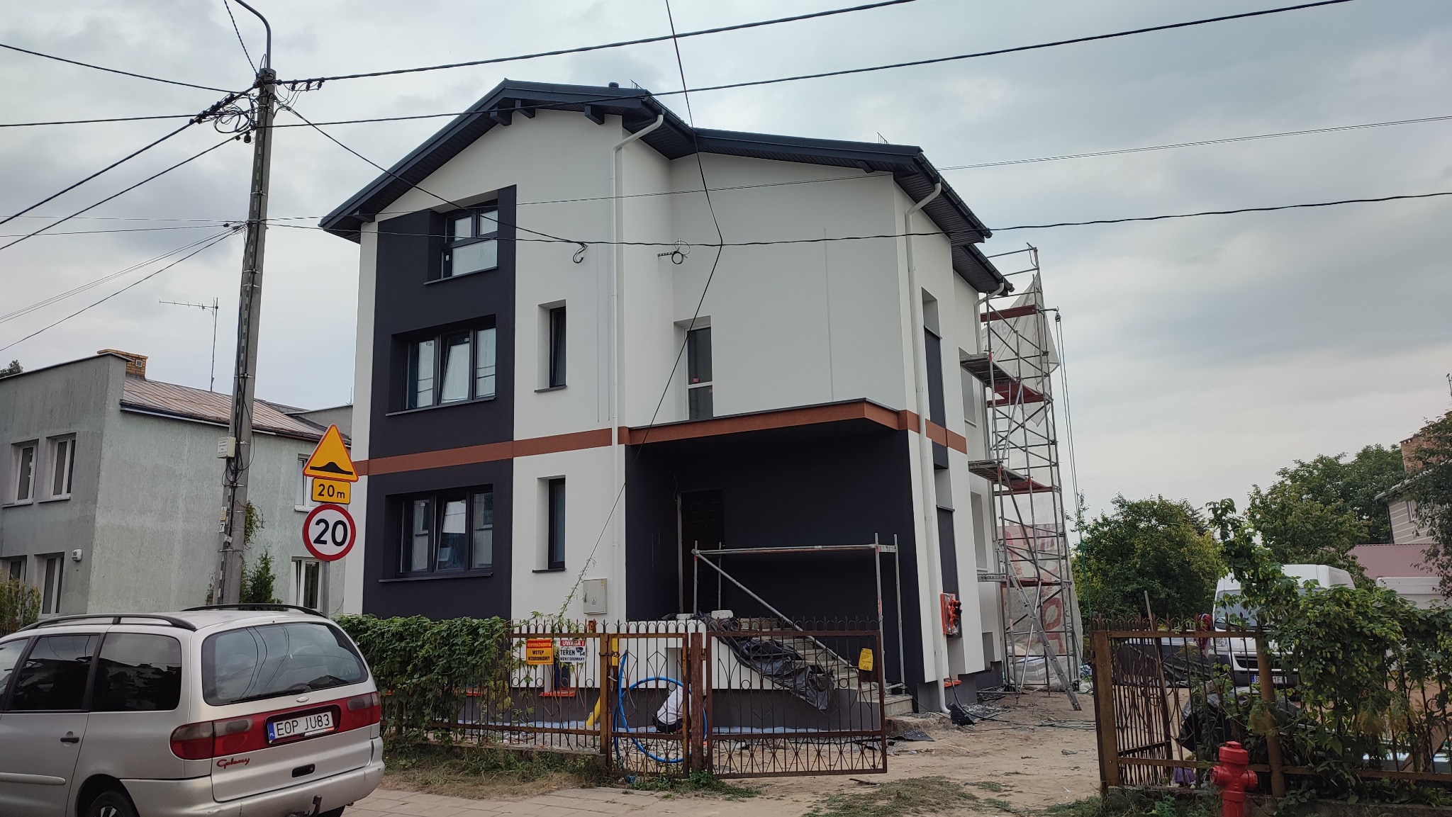 Dwukondygnacyjny dom z elewacją w kolorach białym, czarnym i brązowym, częściowo zasłonięty rusztowaniem, widoczny znak drogowy i samochód w tle.