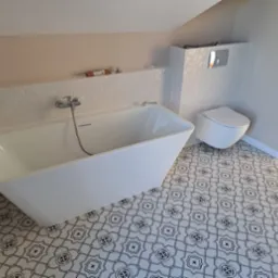 Nowoczesna łazienka z wolnostojącą białą wanną, podwieszanym WC, białą mozaiką na ścianie i wzorzystą czarno-białą podłogą, z widocznym fragmentem beżowej ściany pod skosem.