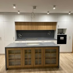 ARCHITON SPÓŁKA Z OGRANICZONĄ ODPOWIEDZIALNOŚCIĄ - Nowoczesna kuchnia z wyspą: białe szafki górne, drewniane dolne z przeszkleniami, szary blat, oświetlenie punktowe. Minimalistyczny design, jasne wnętrze.