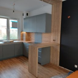 ARCHITON SPÓŁKA Z OGRANICZONĄ ODPOWIEDZIALNOŚCIĄ - Szara kuchnia z drewnianym blatem i barkiem, minimalistyczny design, drewniana podłoga, czarna ściana w tle, surowe wykończenie, widok na okno.