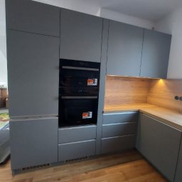ARCHITON SPÓŁKA Z OGRANICZONĄ ODPOWIEDZIALNOŚCIĄ - Szara kuchnia na wymiar z drewnianym blatem i dwoma piekarnikami, oświetlona od spodu, na parkiecie. Minimalistyczny design.