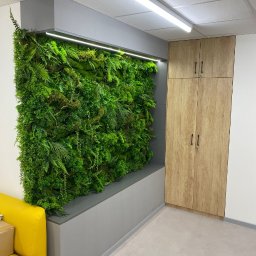 ARCHITON SPÓŁKA Z OGRANICZONĄ ODPOWIEDZIALNOŚCIĄ - Wnętrze z pionową ścianą z roślin i szafą z drewnianymi frontami. Żywa zieleń kontrastuje z szarością zabudowy i podłogi. Fragment żółtej sofy w lewym dolnym rogu.