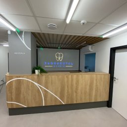 ARCHITON SPÓŁKA Z OGRANICZONĄ ODPOWIEDZIALNOŚCIĄ - Drewniana lada recepcyjna z białymi zdobieniami w Krępicach. Nowoczesny design, oświetlenie LED i minimalistyczny styl. Widok na logo kliniki dentystycznej.