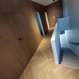 ARCHITON SPÓŁKA Z OGRANICZONĄ ODPOWIEDZIALNOŚCIĄ - Nowoczesny korytarz z ukrytymi drzwiami w panelach ściennych o drewnianym wykończeniu, parkiet w jodełkę i schody w tle. Minimalistyczny design.