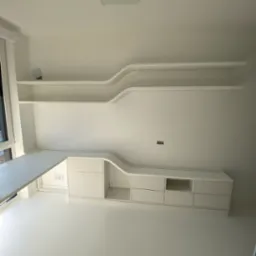 Białe, minimalistyczne meble na wymiar z półkami o falistym kształcie, wkomponowane w jasne wnętrze. Biurko przylega do okna, a całość tworzy spójną, nowoczesną przestrzeń.