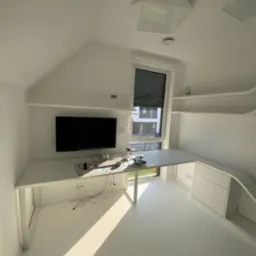 Białe biurko na wymiar w pokoju na poddaszu, zintegrowane z szafkami i półkami. Minimalistyczny design, nowoczesne oświetlenie sufitowe, roleta w oknie.