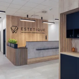 Recepcja w nowoczesnej klinice stomatologicznej z drewnianymi panelami, szarą ladą i elementami zabudowy meblowej w kolorze granatowym, w tle widoczny korytarz z drzwiami.