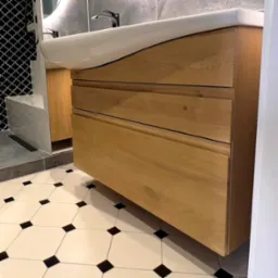 Łazienka z drewnianą szafką pod umywalką z trzema szufladami, białą umywalką z zakrzywionym frontem, mozaiką na ścianie i wzorzystą podłogą w biało-czarne kwadraty.