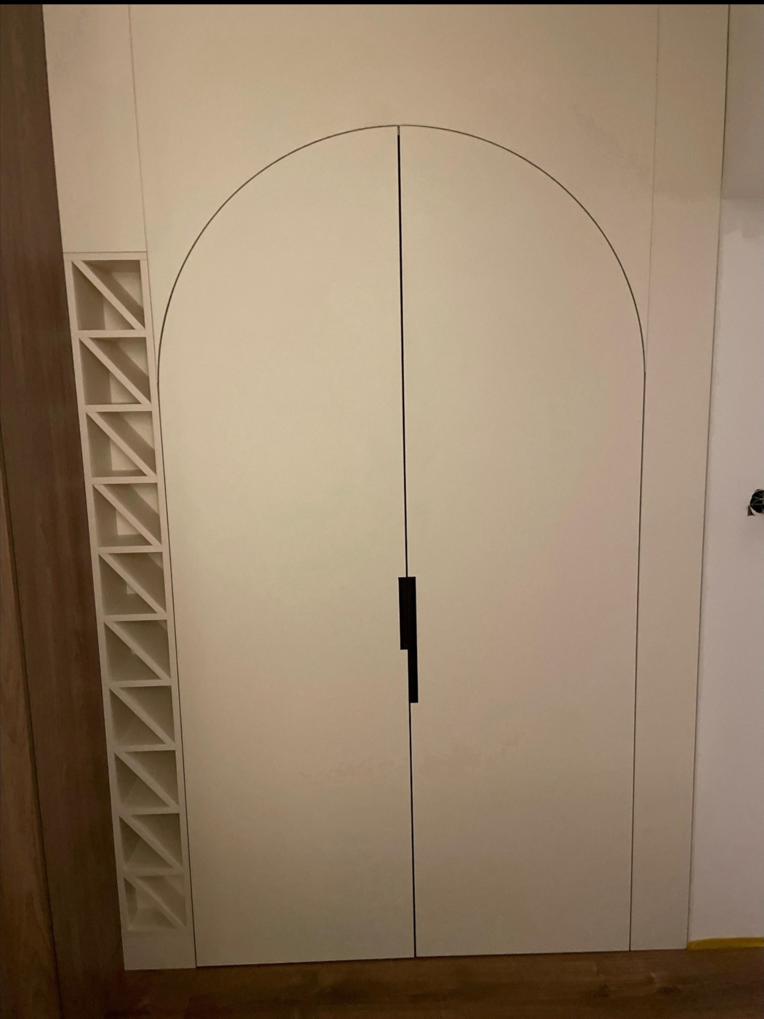 Biała szafa wnękowa z łukowatymi drzwiami i geometrycznym regałem po lewej stronie, na tle drewnianej podłogi. Minimalistyczny design.