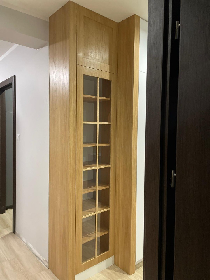 Drewniana szafa z przeszklonymi drzwiczkami, wnęki z półkami, jasne drewno, widok z boku, minimalistyczny design, w tle biała ściana i ciemne drzwi.