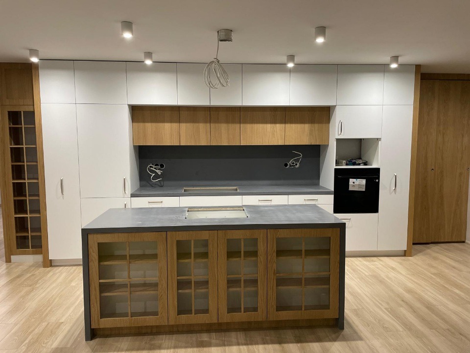 Nowoczesna kuchnia z wyspą: białe szafki górne, drewniane dolne z przeszkleniami, szary blat, oświetlenie punktowe. Minimalistyczny design, jasne wnętrze.