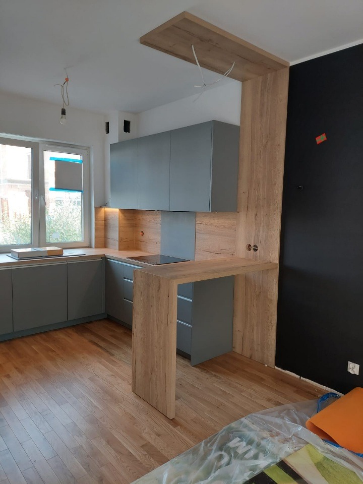 Szara kuchnia z drewnianym blatem i barkiem, minimalistyczny design, drewniana podłoga, czarna ściana w tle, surowe wykończenie, widok na okno.