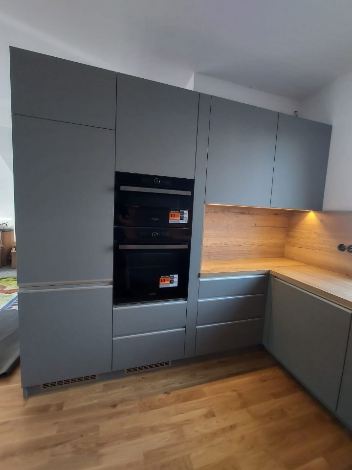 Szara kuchnia na wymiar z drewnianym blatem i dwoma piekarnikami, oświetlona od spodu, na parkiecie. Minimalistyczny design.