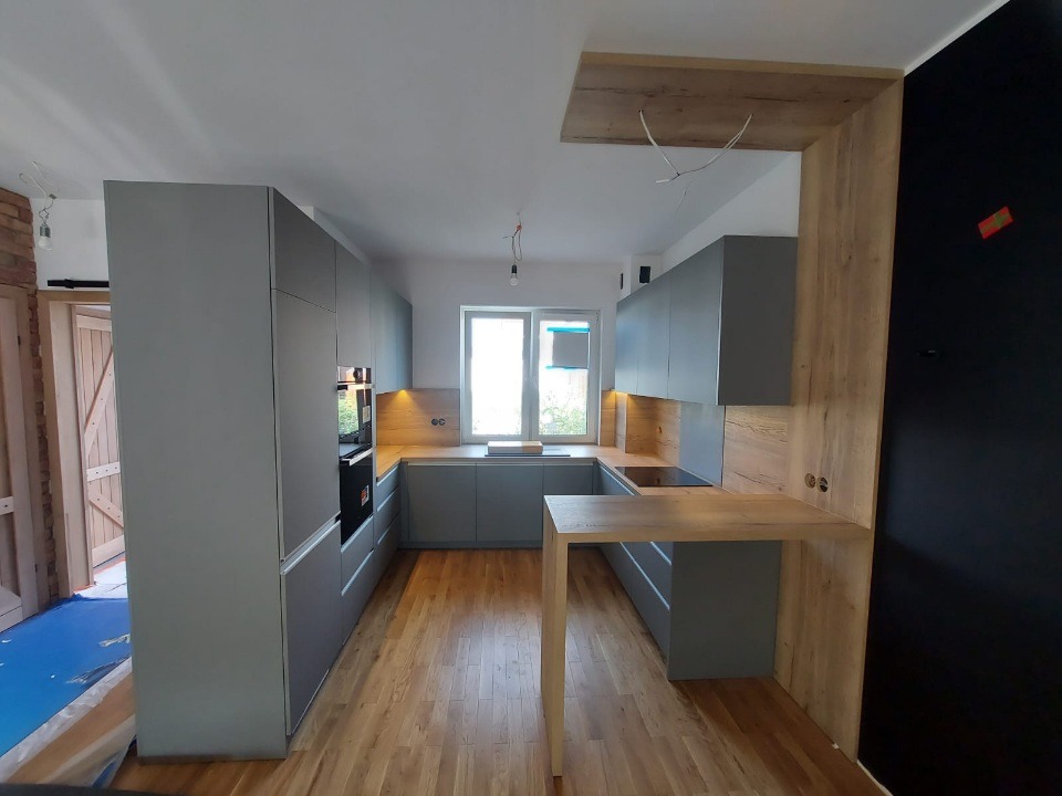 Nowoczesna kuchnia w szarościach z drewnianymi akcentami: zabudowa kuchenna, wyspa z blatem, oświetlenie podszafkowe, parkiet. Minimalistyczny design z elementami naturalnego drewna.