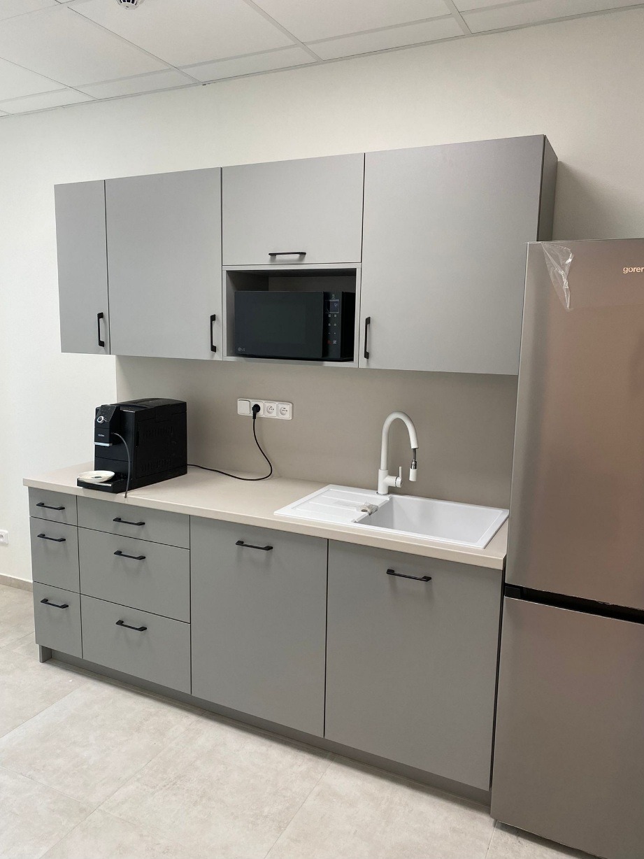 Szara zabudowa kuchenna z czarnymi uchwytami, zintegrowana z mikrofalówką i ekspresem do kawy, uzupełniona o biały zlew i srebrną lodówkę.