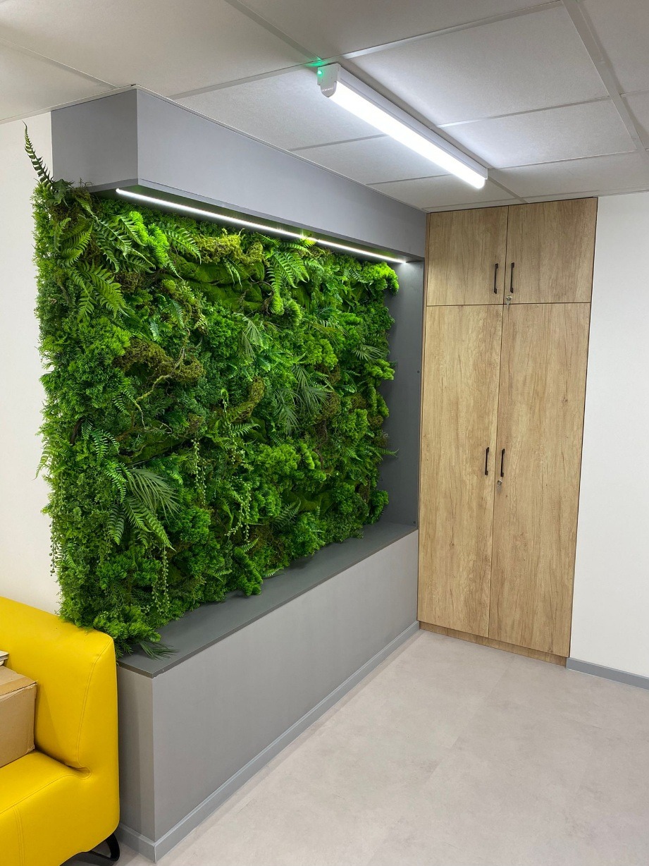 Wnętrze z pionową ścianą z roślin i szafą z drewnianymi frontami. Żywa zieleń kontrastuje z szarością zabudowy i podłogi. Fragment żółtej sofy w lewym dolnym rogu.