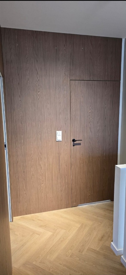 Drewniana zabudowa ściany z ukrytymi drzwiami i włącznikiem światła. Parkiet ułożony w jodełkę. Minimalistyczny design.