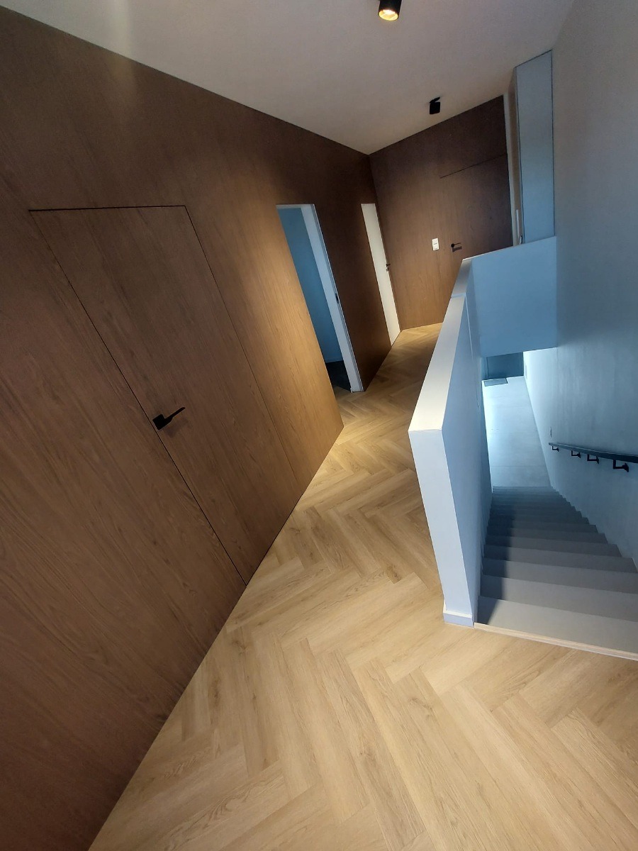 Nowoczesny korytarz z ukrytymi drzwiami w panelach ściennych o drewnianym wykończeniu, parkiet w jodełkę i schody w tle. Minimalistyczny design.