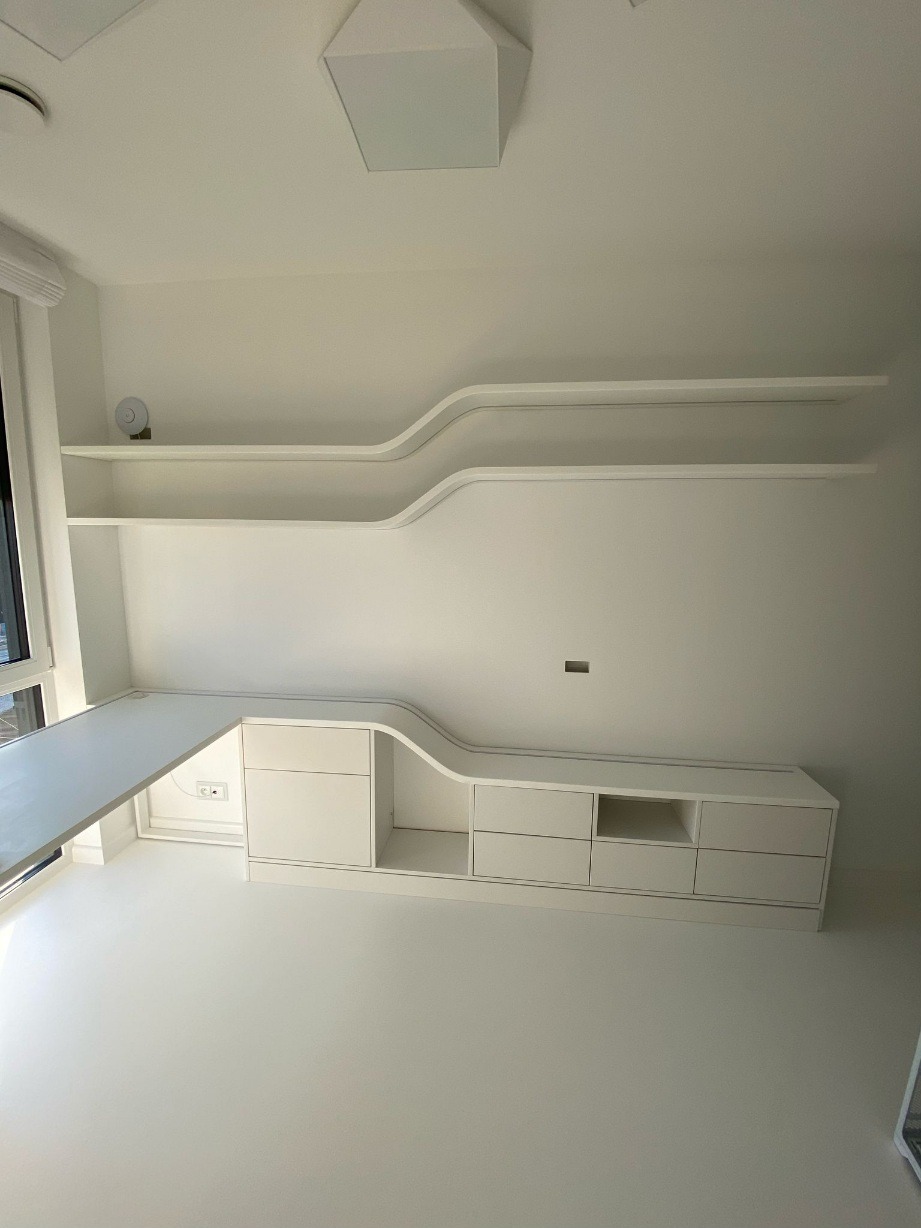 Białe, minimalistyczne meble na wymiar z półkami o falistym kształcie, wkomponowane w jasne wnętrze. Biurko przylega do okna, a całość tworzy spójną, nowoczesną przestrzeń.