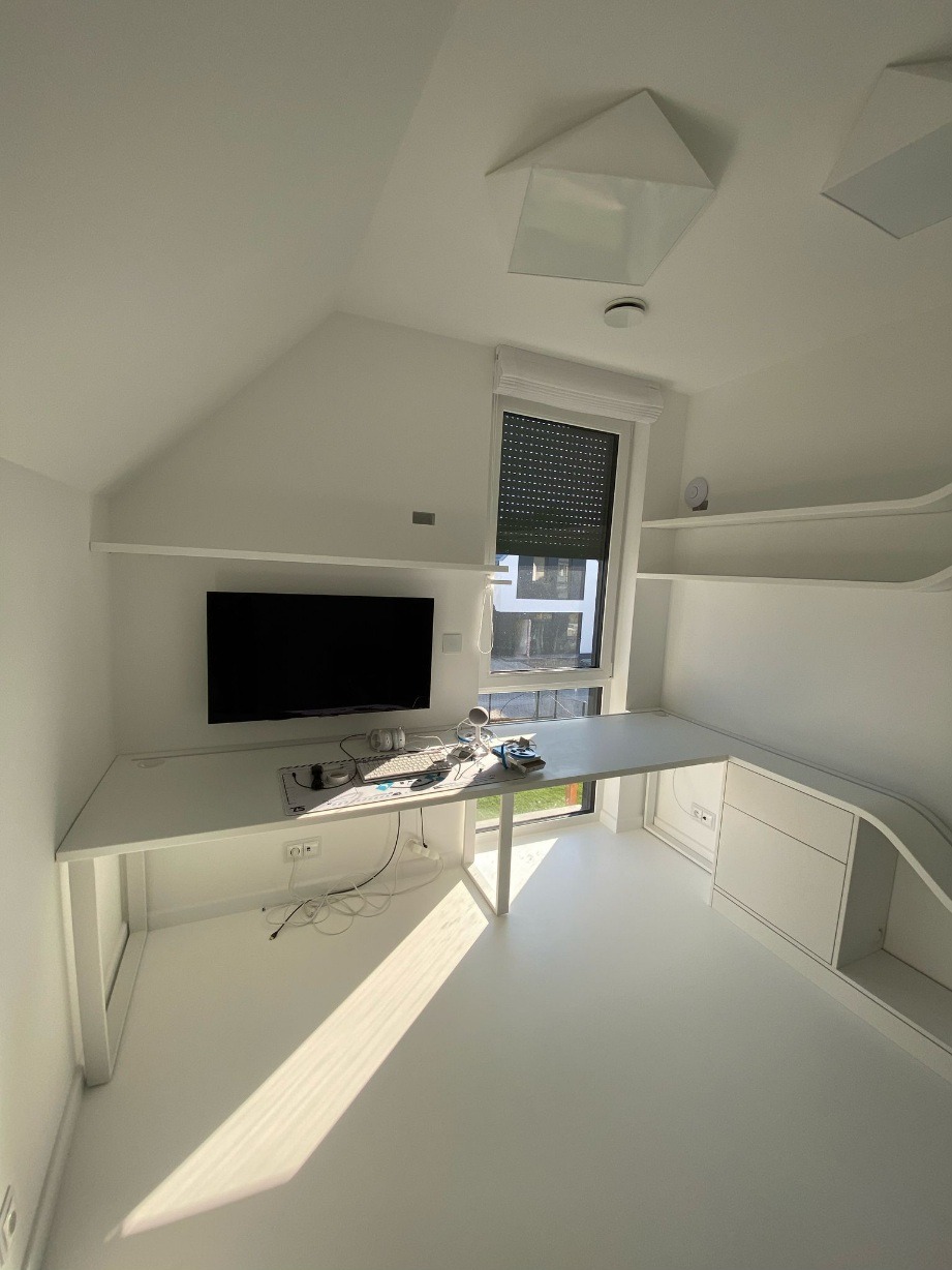 Białe biurko na wymiar w pokoju na poddaszu, zintegrowane z szafkami i półkami. Minimalistyczny design, nowoczesne oświetlenie sufitowe, roleta w oknie.