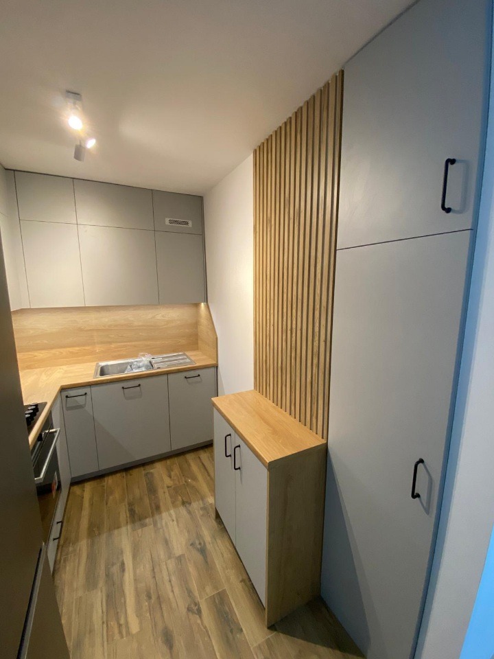 Jasna kuchnia z drewnianymi akcentami: lamele na ścianie i blat. Szare szafki z czarnymi uchwytami, drewnopodobna podłoga. Nowoczesny design, minimalistyczny styl.