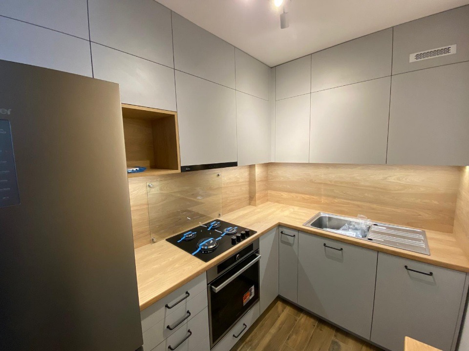 Szara kuchnia z drewnianym blatem i szklanym panelem nad płytą grzewczą. Minimalistyczny design, oświetlenie LED pod szafkami. Nowoczesne wykończenie.
