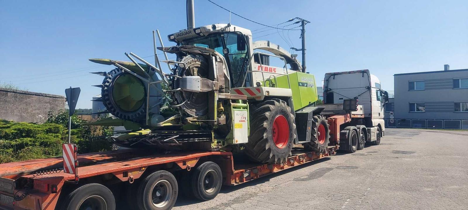 Zielono-biały kombajn rolniczy Jaguar 870 na niskopodwoziowej naczepie transportowany przez biały ciągnik siodłowy. Naczepa z czerwonymi elementami mocującymi. Na pierwszym planie zaparkowany...