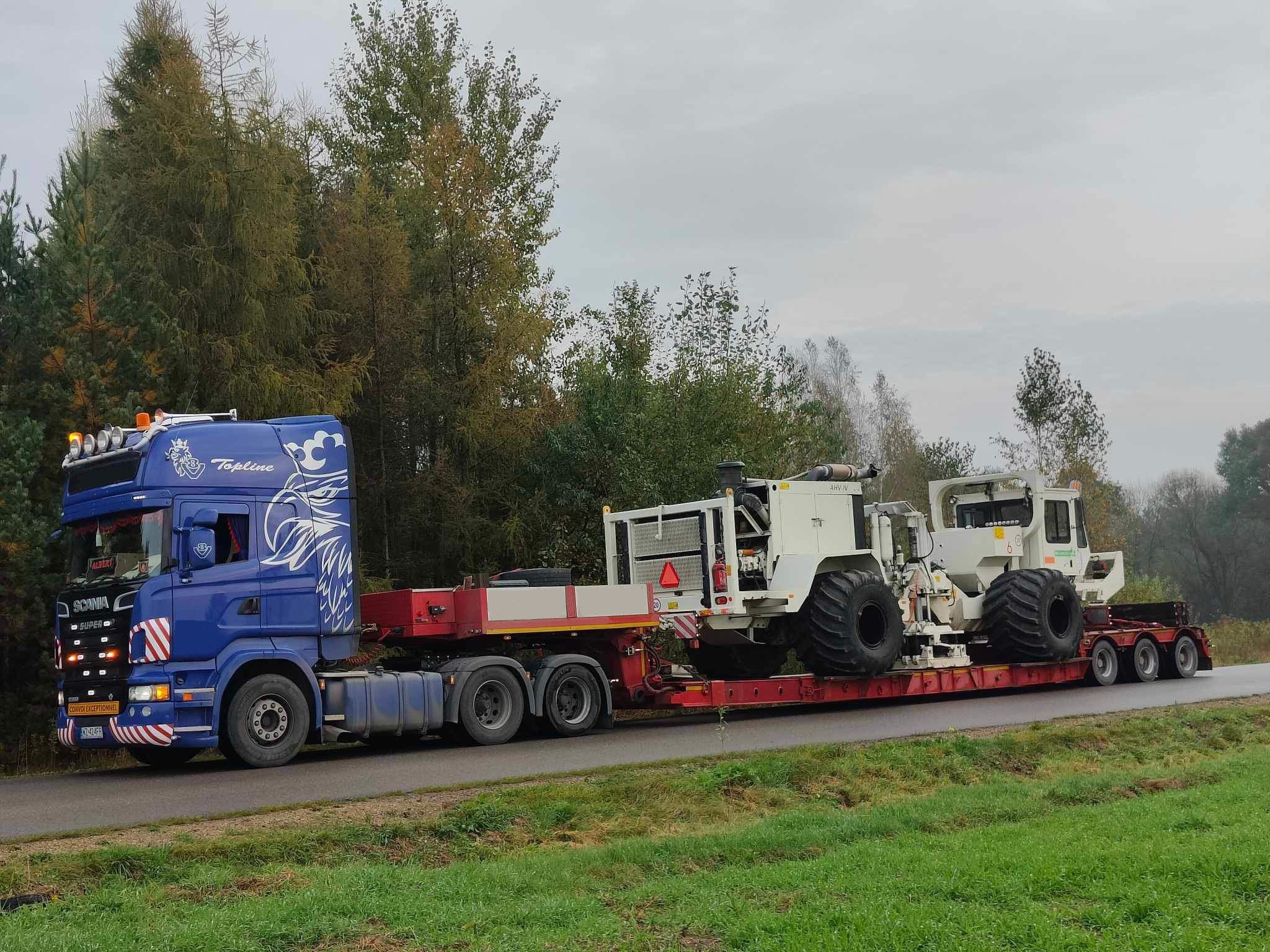 Niebieska ciężarówka Scania z grafiką orła na kabinie przewozi na niskopodwoziowej platformie dwa białe pojazdy specjalistyczne z dużymi oponami, widok z drogi w okolicach Łodzi.