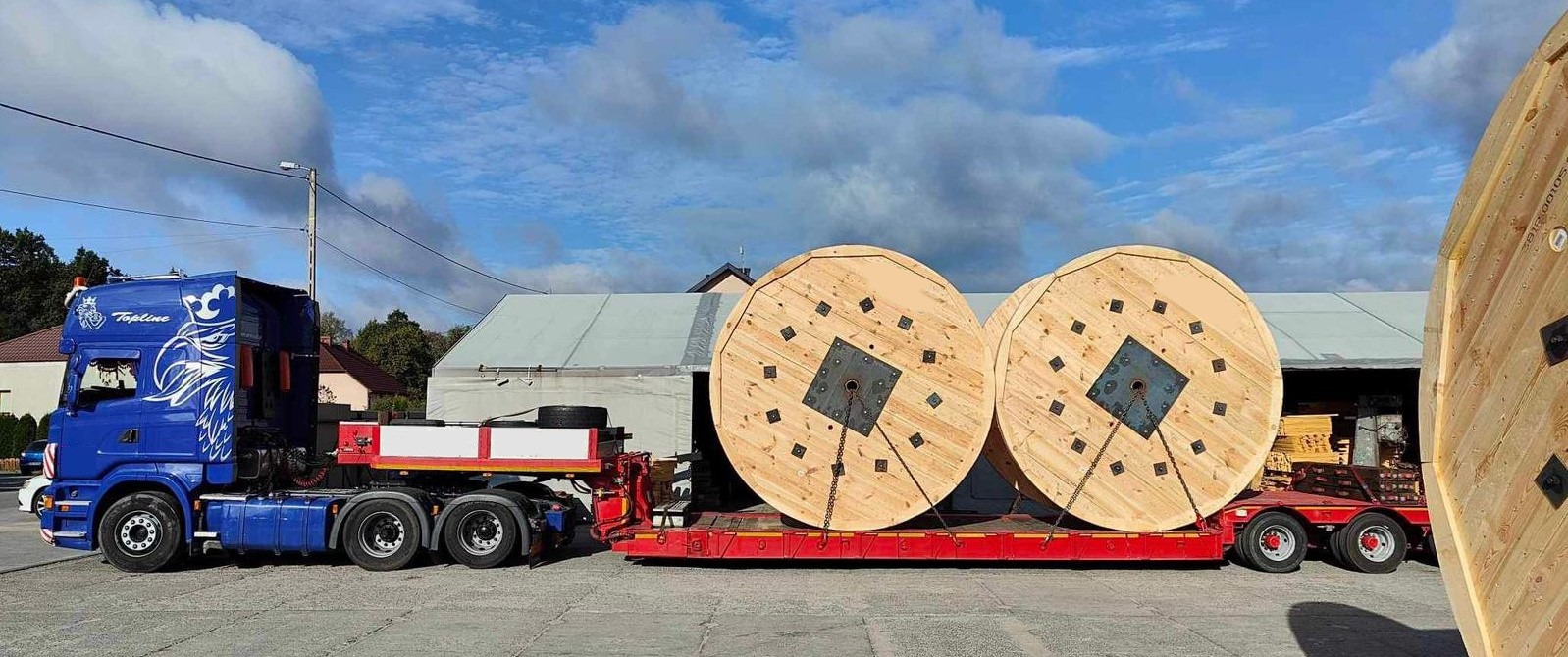 Niebieska ciężarówka z grafiką orła na kabinie transportuje na czerwonej platformie dwa ogromne, drewniane bębny na kable, zabezpieczone łańcuchami.